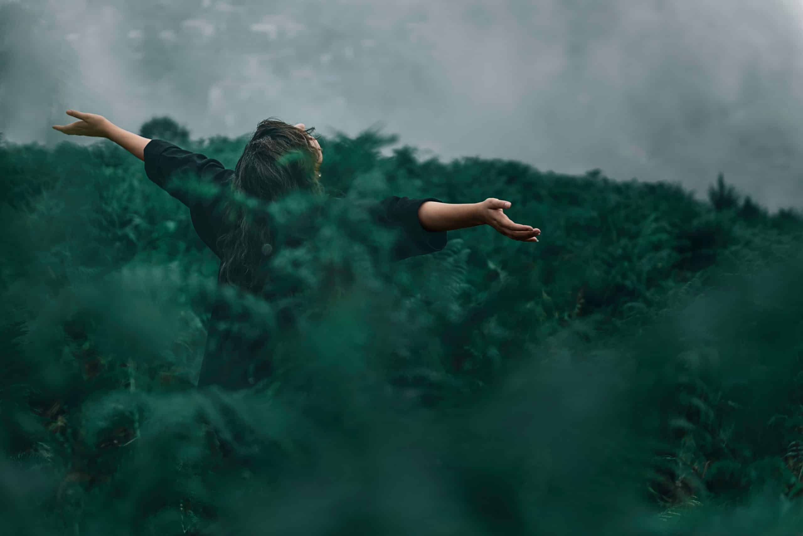 woman tips head back and stretches arms out in the middle of a forest